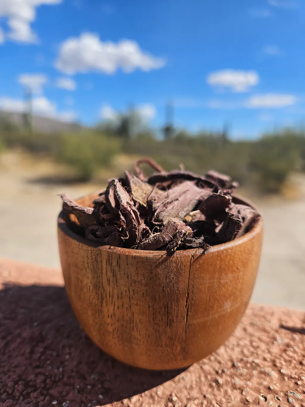 A wooden bowl filled with dark, irregular Mimosa hostilis root bark chips outdoors.